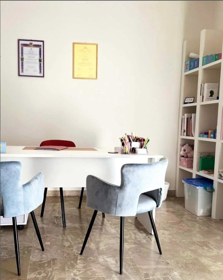 Lo studio della Dott.ssa Gentile: scrivania bianca, poltroncine azzurre, libreria con materiali per bambini e i diplomi alla parete